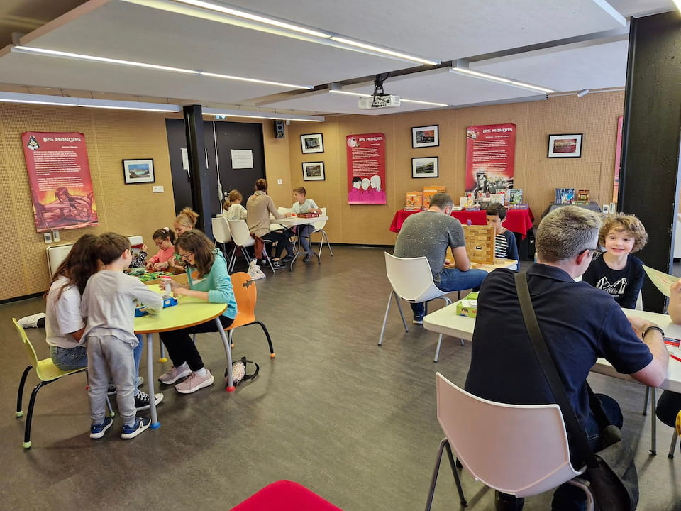 Enfants et adultes assis autour de tables dans une salle, et jouant à des jeux de société